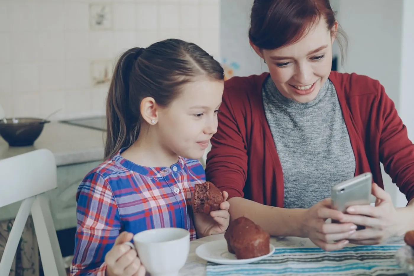 Una madre y su hija miran juntas el celular en la cocina